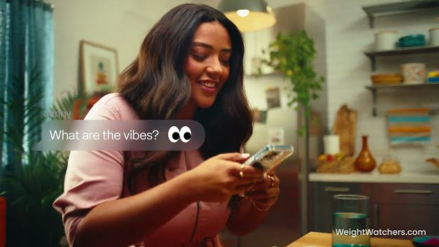 Happy woman in a pink shirt using smartphone in a bright kitchen setting with WeightWatchers.com branding