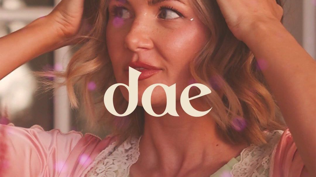 A woman with sun-kissed waves and a gleeful expression is adorned with twinkling purple orbs and the elegant "dae" logo, evoking a sense of magical haircare charm.