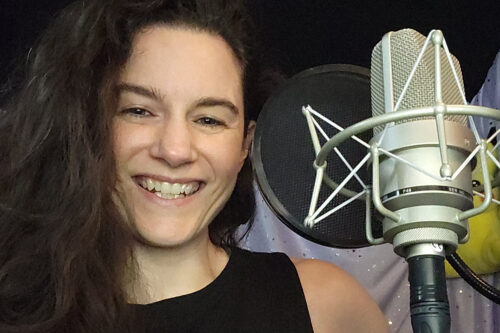 Voice actor Nina Nikolic smiling in her home recording studio, with a large diaphragm condenser microphone in shock mount positioned beside her. She wears a black sleeveless top against a backdrop of acoustic treatment panels, capturing the professional setup of a sustainable freelance business in voice acting.