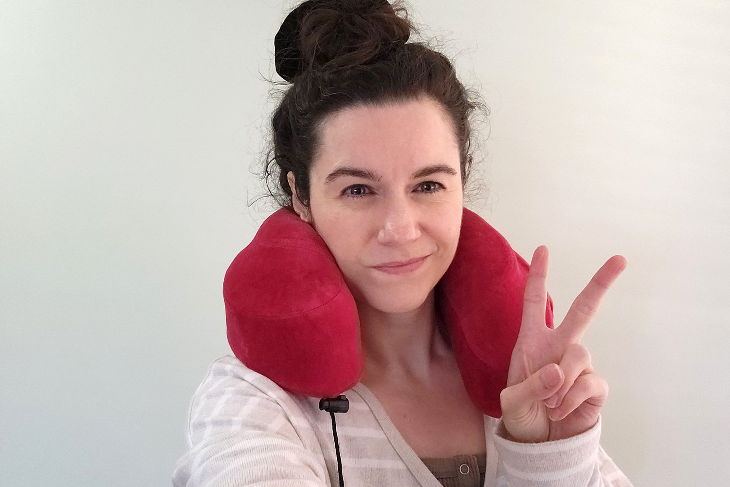 Nina Nikolic wearing a red travel neck pillow and casual loungewear, flashing a peace sign while smiling at the camera. The travel pillow represents the bi-city lifestyle between Melbourne and Sydney that became essential for balancing freelance work with family caregiving priorities.
